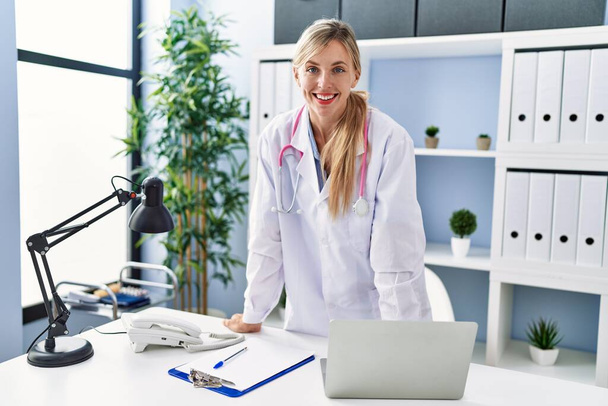 Jonge blonde vrouw draagt dokter uniform staan bij kliniek - Foto, afbeelding