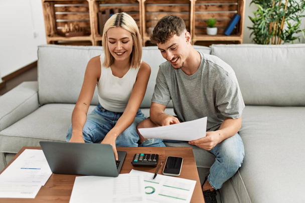 Młoda kaukaska para uśmiechnięta szczęśliwa kontrolując gospodarkę rodzinną za pomocą laptopa w domu. - Zdjęcie, obraz