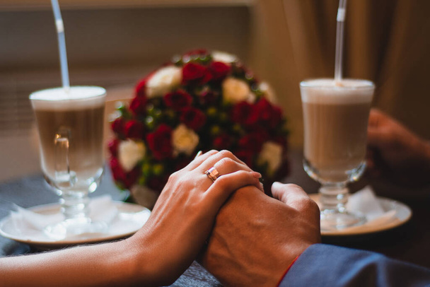 Liebendes Brautpaar, das sich Händchen haltend auf dem Hintergrund von zwei Gläsern Cappuccino und einem Brautstrauß aus roten und weißen Blumen nähert. Hochzeitsfoto in einem gemütlichen Café am Hochzeitstag. - Foto, Bild