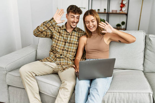 Молодая латиноамериканская пара с видеозвонком с помощью ноутбука сидит на диване дома. - Фото, изображение