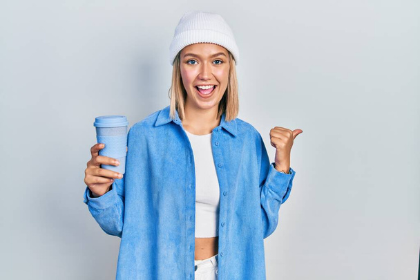 Beautiful blonde woman drinking a take away cup of coffee pointing thumb up to the side smiling happy with open mouth  - Фото, изображение