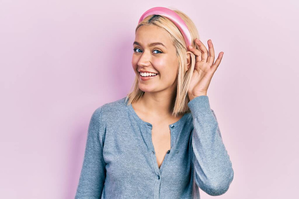 Beautiful blonde woman standing over pink background smiling with hand over ear listening an hearing to rumor or gossip. deafness concept.  - Fotografie, Obrázek