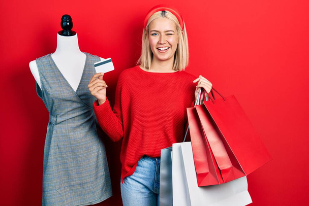 Schöne blonde Frau mit Einkaufstaschen und Kreditkarte zwinkernd in die Kamera blickend mit sexy Gesichtsausdruck, fröhlichem und glücklichem Gesicht.  - Foto, Bild