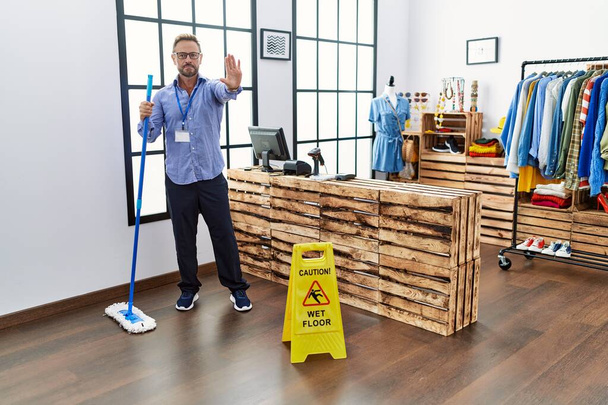 Středního věku muž pracující v maloobchodě úklid podlahy s otevřenou rukou dělat stop sign s vážným a sebevědomým výrazem, obranné gesto  - Fotografie, Obrázek