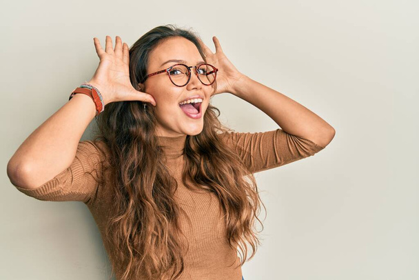 Νεαρό ισπανόφωνο κορίτσι που φοράει καθημερινά ρούχα και γυαλιά χαμογελώντας χαρούμενα παίζοντας κρυφοκοιτάζει ένα boo με τα χέρια που δείχνουν το πρόσωπο. έκπληκτος και εξερχόμενος  - Φωτογραφία, εικόνα