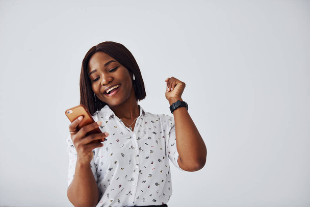 Positive african american woman in white shirt standing indoors against white wall with phone in hand. - Fotografie, Obrázek