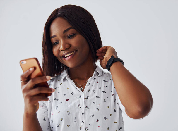 Positieve Afro-Amerikaanse vrouw in wit shirt staat binnen tegen een witte muur met telefoon in de hand. - Foto, afbeelding