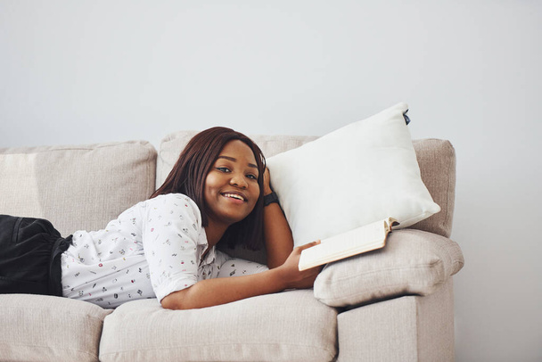 Positive african american woman in white shirt lying down on sofa with notepad in hands. - Foto, imagen
