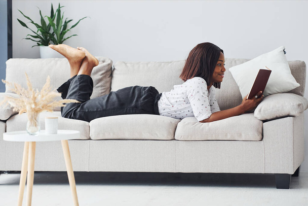 Positive african american woman in white shirt lying down on sofa with notepad in hands. - Foto, immagini
