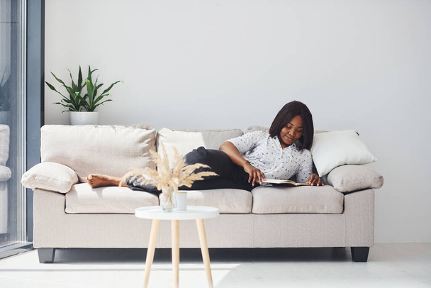 Positive african american woman in white shirt lying down on sofa with notepad in hands. - Foto, afbeelding