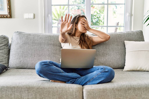 Piękna młoda brunetka siedzi na kanapie za pomocą laptopa komputerowego w domu przykrywając oczy rękami i wykonując stop gest z smutku i strachu ekspresji. wstydliwa i negatywna koncepcja.  - Zdjęcie, obraz