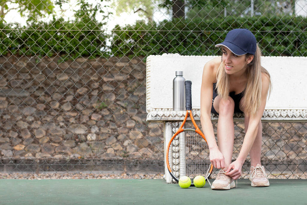 Портрет тенісистки, що прив'язує взуття, сидить на лавці в тенісному корті в шапці з копіювальним простором
 - Фото, зображення