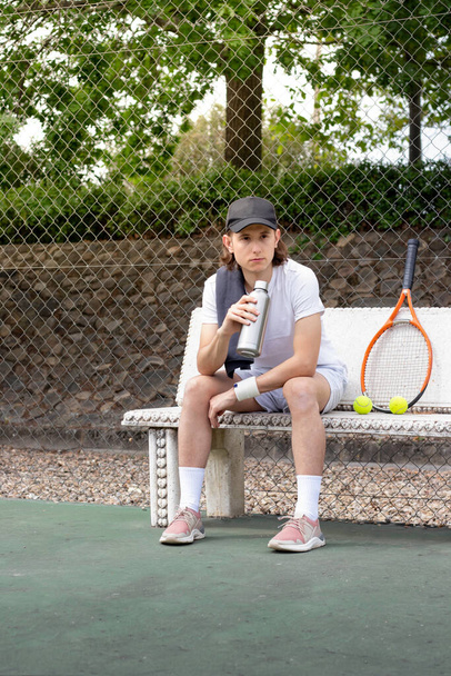 Joven descansando sentado en un banco después de jugar un partido de tenis con una botella de agua en la mano y una raqueta - Foto, Imagen