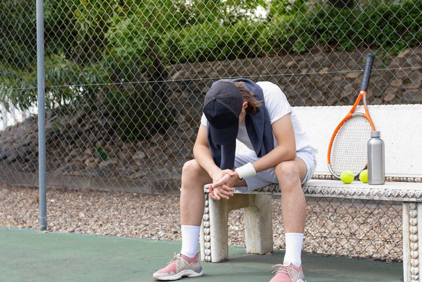 Εξαντλημένος αθλητής κάθεται σε ένα παγκάκι μετά το μάθημα τένις κοιτάζοντας προς τα κάτω στο πάτωμα στο δικαστήριο - Φωτογραφία, εικόνα