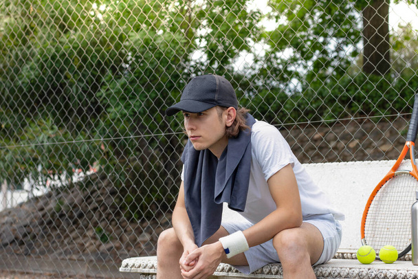 Πορτρέτο του νεαρού άνδρα συγκεντρωμένη ψάχνει σε έναν αγώνα τένις, ενώ κάθεται σε ένα παγκάκι με μια πετσέτα γύρω από το λαιμό του - Φωτογραφία, εικόνα
