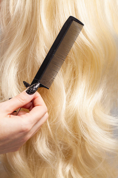 Friseur hält Kamm auf langen lockigen blonden Haaren Hintergrund Nahaufnahme - Foto, Bild