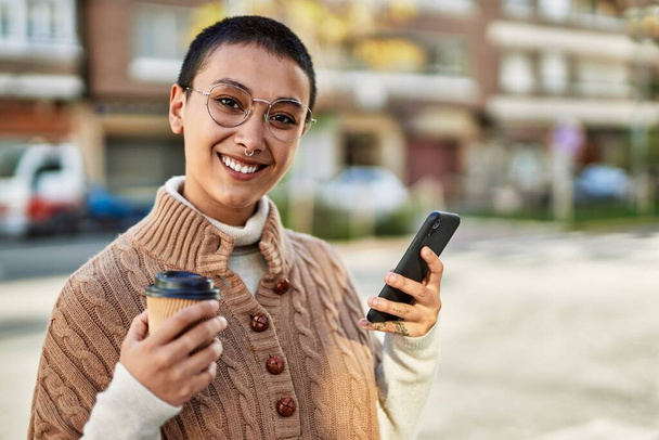 Jonge Spaanse vrouw met kort haar glimlachend gelukkig drinken van een kopje koffie en met behulp van smartphone - Foto, afbeelding