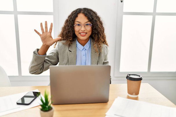 Belle femme d'affaires hispanique assis sur le bureau au bureau travaillant avec ordinateur portable montrant et pointant vers le haut avec les doigts numéro cinq tout en souriant confiant et heureux.  - Photo, image