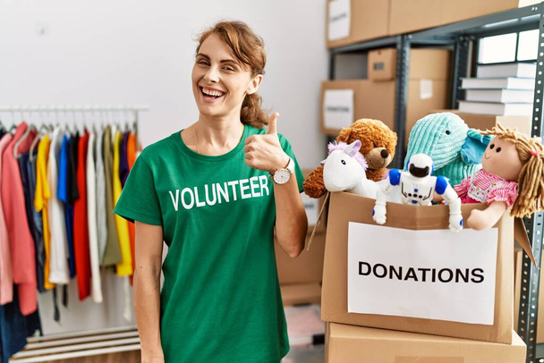 Beautiful caucasian woman wearing volunteer t shirt at donations stand doing happy thumbs up gesture with hand. approving expression looking at the camera showing success.  - Photo, Image
