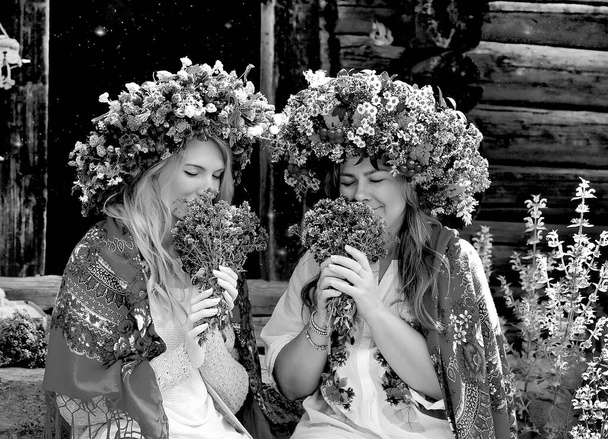 Schwarz-weiße Mädchen in großen Blumenkränzen, ukrainische Nationalschals auf den Schultern, schnüffelnde Blumensträuße. Hintergrund Holzhaus. Sommer. Traditionen in der Ukraine - Foto, Bild