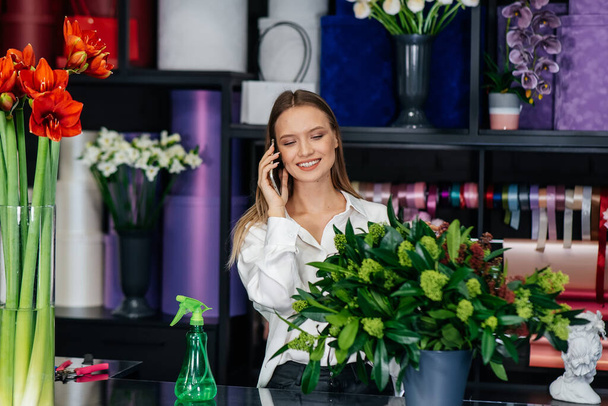 A young woman florist accepts an order by phone for the production of a beautiful festive bouquet. Floristry and making of bukets in a flower shop. Small business. - 写真・画像