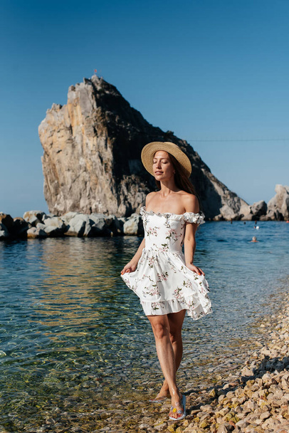 A beautiful young woman in a hat, glasses and a light dress is walking along the ocean shore against the background of huge rocks on a sunny day. Tourism and tourist trips. - Valokuva, kuva