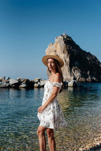 A beautiful young woman in a hat, glasses and a light dress is walking along the ocean shore against the background of huge rocks on a sunny day. Tourism and tourist trips. - Фото, изображение