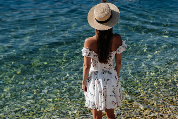 A beautiful young woman in a hat and a light dress with her back is walking along the ocean shore against the background of huge rocks on a sunny day. Tourism and vacation travel. - Фото, изображение