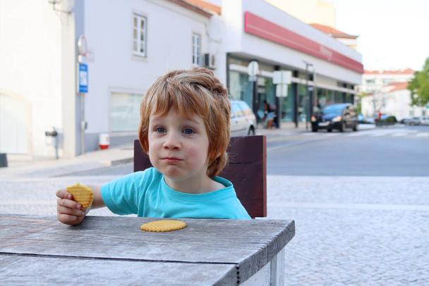 Um menino loiro senta-se em uma mesa de madeira em um café de rua comendo biscoitos. O menino de cinco anos está comendo e sorrindo. - Foto, Imagem