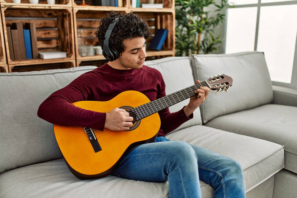 Młody Latynos gra na gitarze klasycznej siedząc na kanapie w domu. - Zdjęcie, obraz
