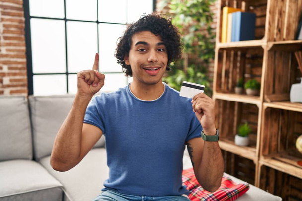 Homme hispanique avec les cheveux bouclés tenant la carte de crédit surpris par une idée ou une question pointant du doigt avec le visage heureux, numéro un  - Photo, image