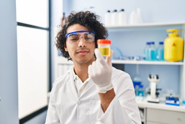 Joven hispano vistiendo uniforme científico usando portátil analizando sonda de orina en laboratorio - Foto, Imagen
