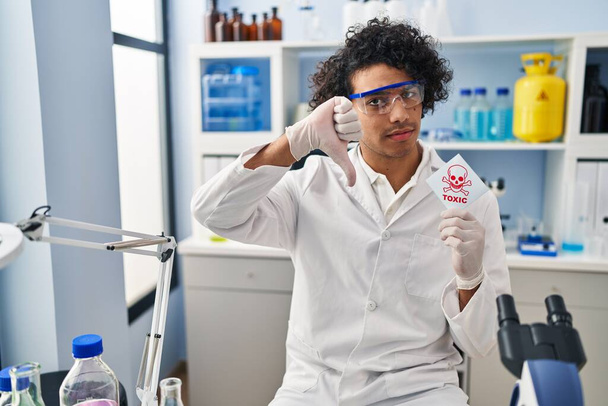 Hispanic man with curly hair working at scientist laboratory holding toxic banner with angry face, negative sign showing dislike with thumbs down, rejection concept  - Foto, immagini
