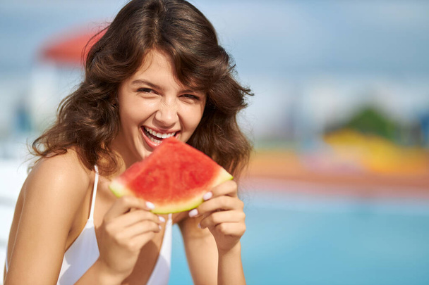 Happy wavy haired female smiling in pleasure, while biting watermelon outside. - Foto, afbeelding