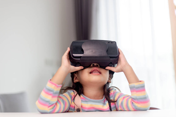 Enfant avec casque de réalité virtuelle assis derrière la table à l'intérieur à la maison - Photo, image