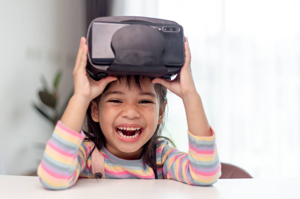 Criança com fone de ouvido de realidade virtual sentada atrás da mesa dentro de casa - Foto, Imagem