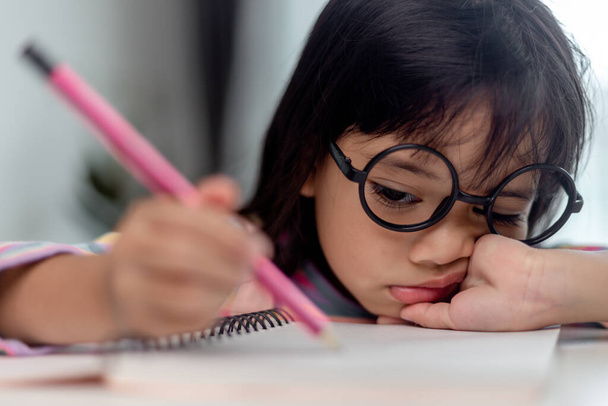 Klein Aziatisch meisje zitten alleen en kijken uit met een verveeld gezicht, kleuter liggend hoofd neer op de tafel met trieste verveeld met huiswerk, verwend kind - Foto, afbeelding
