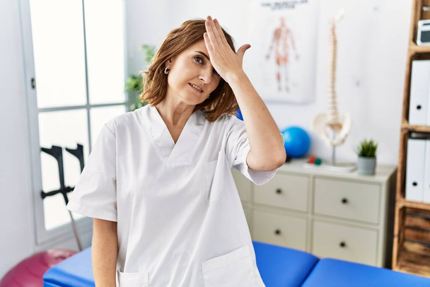 Middle age physiotherapist woman working at pain recovery clinic surprised with hand on head for mistake, remember error. forgot, bad memory concept.  - Photo, Image