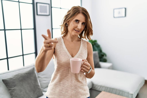 Keski-ikäinen nainen juo kuppi kahvia kotona osoittaa sormella kameraan onnellinen ja hauska kasvot. hyvä energia ja vibes.  - Valokuva, kuva