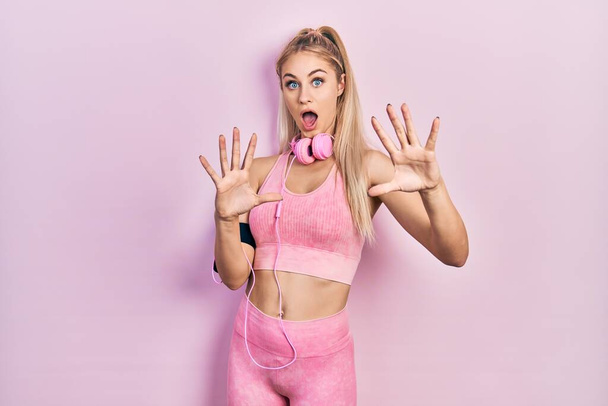 Young beautiful caucasian woman wearing gym clothes and using headphones afraid and terrified with fear expression stop gesture with hands, shouting in shock. panic concept.  - Photo, Image