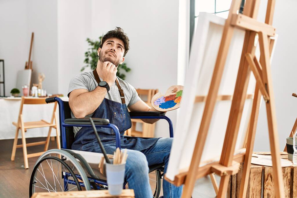 Joven hombre hispano sentado en silla de ruedas pintando en el estudio de arte pensando concentrado en la duda con el dedo en la barbilla y mirando hacia arriba preguntándose  - Foto, imagen