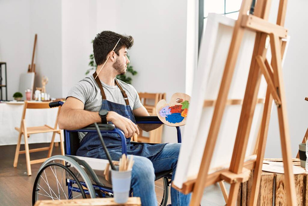 Joven hombre hispano sentado en la pintura en silla de ruedas en el estudio de arte mirando al costado, pose de perfil relajado con rostro natural y sonrisa confiada.  - Foto, Imagen