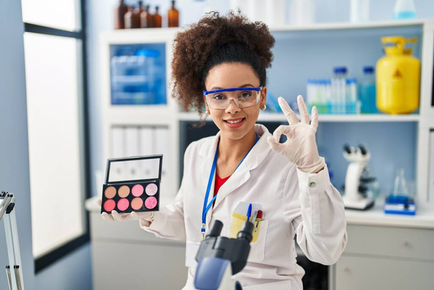 Młoda Afroamerykanka pracująca w laboratorium robi makijaż robiąc znak ok palcami, uśmiechając się przyjazny gest doskonały symbol  - Zdjęcie, obraz