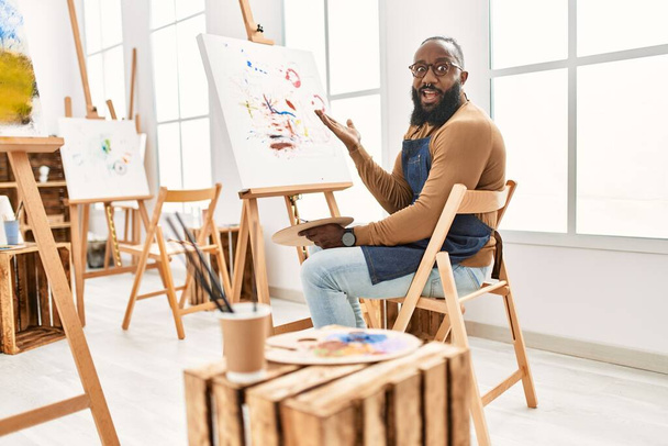 アフリカ系アメリカ人アーティストの男性が笑顔で勝利を祝うアートスタジオでキャンバスに絵を描き、手を上げて勝者の表情  - 写真・画像