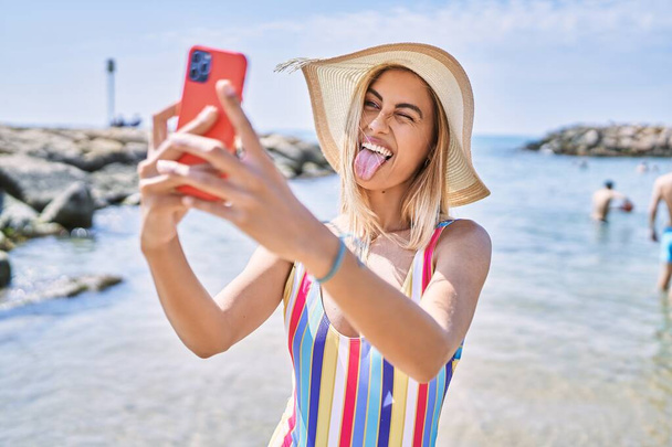 Молода блондинка в літньому капелюсі робить селфі на смартфоні на пляжі
. - Фото, зображення