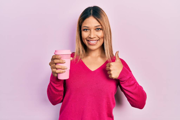 Beautiful hispanic woman drinking a take away cup of coffee smiling happy and positive, thumb up doing excellent and approval sign  - Photo, Image