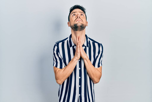 Молодий іспаномовний чоловік в повсякденному одязі молиться і молиться разом з надією висловлюватися на обличчі дуже емоційно і стурбовано. благання
.  - Фото, зображення