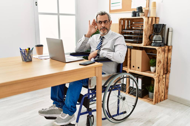 Латиноамериканец средних лет, работающий в офисе, сидя в инвалидном кресле и улыбаясь рукой над ухом, слушает слухи или сплетни. концепция глухоты.  - Фото, изображение