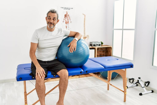 Middle age hispanic man at pain recovery clinic holding pilates ball smiling looking to the side and staring away thinking.  - Photo, Image