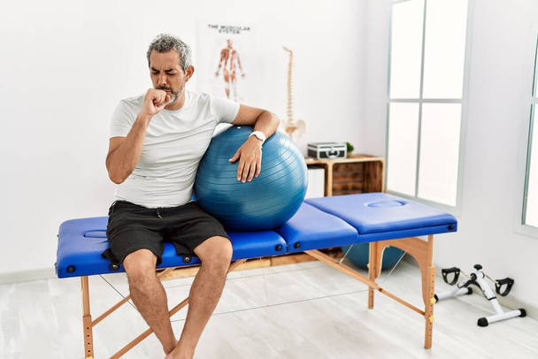 Orta yaşlı İspanyol bir adam ağrı kesici kliniğinde elinde pilates topu tutarken kendini iyi hissetmiyor ve soğuk algınlığı veya bronşitin belirtisi olarak öksürüyor. sağlık hizmeti kavramı.  - Fotoğraf, Görsel